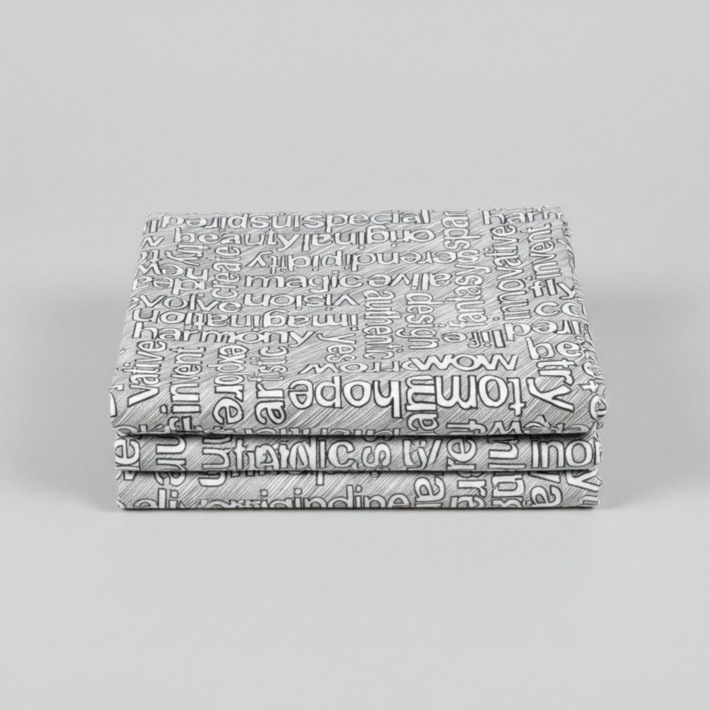 Writing on the Wall Well Worded White quilting cotton by FreeSpirit Fabrics -- three folded yards stacked on a grey surface, grey and white ground with white outlined words in varying sizes and directions including special, beauty, serendipity, magic, imagination, harmony, wow, and tomorrow