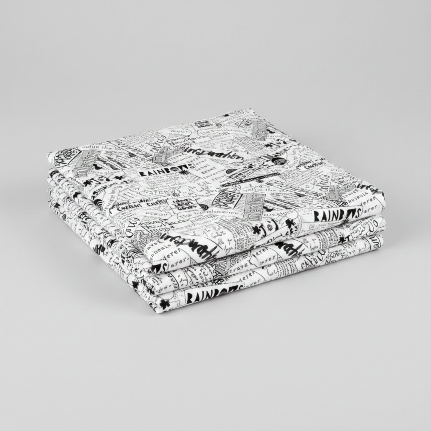 Writing on the Wall Word Collage White quilting cotton by FreeSpirit Fabrics — three folded yards stacked on a grey surface, bold black and white word collage print with overlapping handwritten words including imagination, inspiration, joy, and rainbows