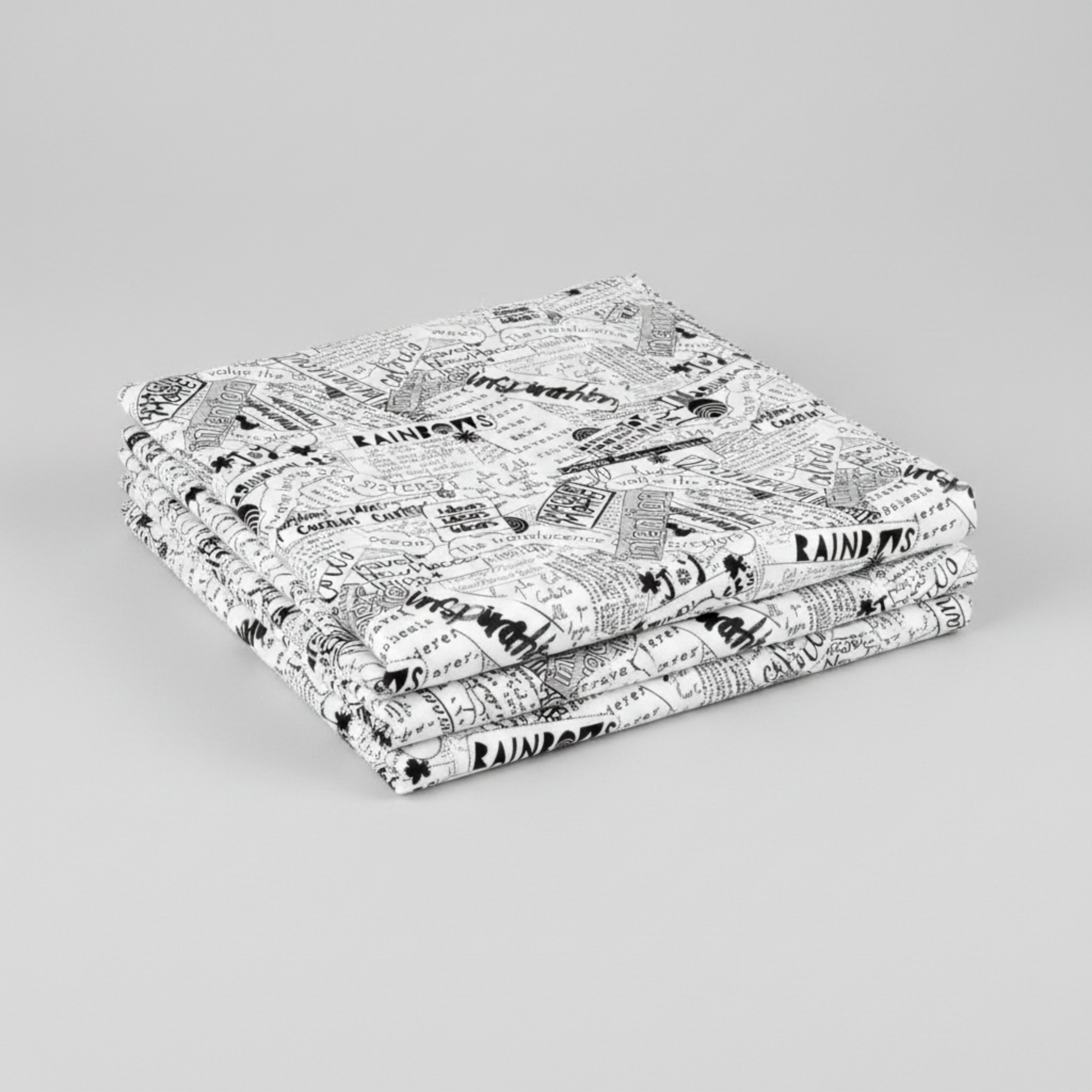 Writing on the Wall Word Collage White quilting cotton by FreeSpirit Fabrics — three folded yards stacked on a grey surface, bold black and white word collage print with overlapping handwritten words including imagination, inspiration, joy, and rainbows